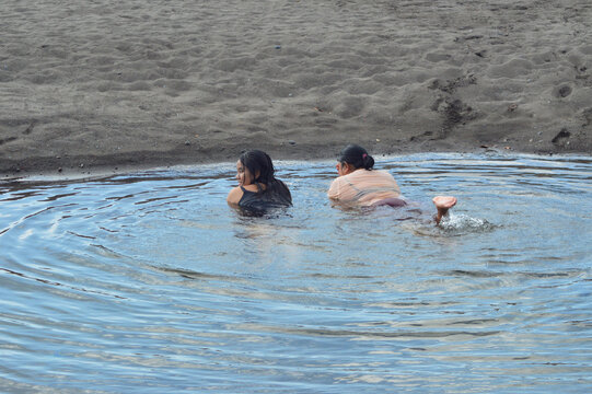 A woman lies prone in shallow sandy creek water, splashing gently with her legs while enjoying relaxation and playful interaction with her daughter nearby.