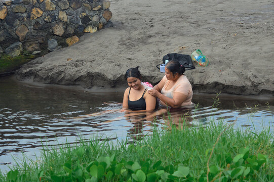 A smiling teenage girl enjoys the moment as her mother washes her back with a shower puff, both seated in calm creek water along the sandy shore.