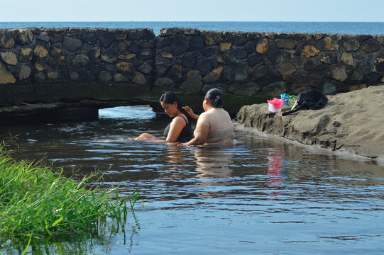 A mother gently combs her teenage daughterâ€™s freshly washed hair as they sit in a shallow brackish creek near the sandy shoreline, enjoying a peaceful beachside moment.