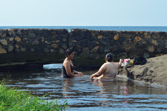 A mother leans forward to pick up bathing items as her daughter waits beside her, both immersed in shallow water near the sandy river mouth under natural daylight.