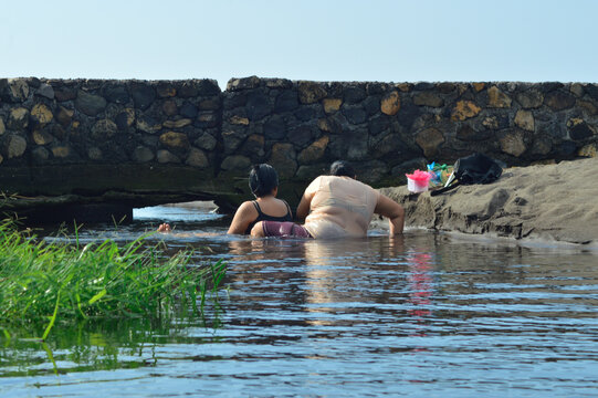 A mother adjusts her seat to reach for bathing supplies while her daughter sits beside her in shallow creek water near the sandy coast under open tropical light.