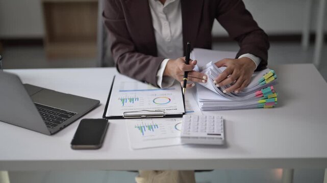 A woman in a business suit is sitting at a desk with a laptop, a cell phone, and a stack of papers. She is writing on a piece of paper with a pen. Concept of productivity and focus