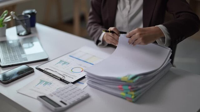A woman is sitting at a desk with a stack of papers in front of her. She is looking at the papers and she is focused on them