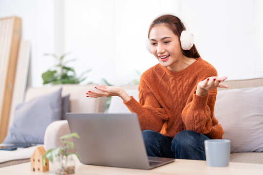 Happy young Asian woman, wearing headphones, is actively engaged in a video call on her laptop, sitting on a sofa and gesticulating with her hands, conveying an expressive and lively conversation.