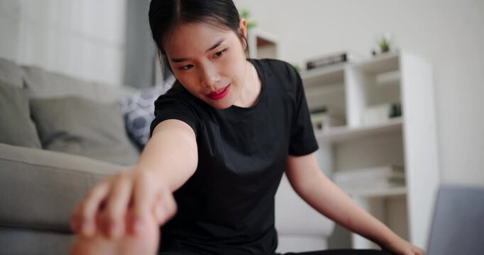 Handheld selective focus shot, Young Asian woman practicing stretching online on laptop in living room at home. Healthy lifestyle. Women Lifestyle