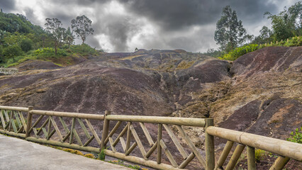 Fototapeta premium A unique natural landmark, 23 coloured earth. Colorful sand hills. Green vegetation on the mountainside. Wooden railings along the footpath. Clouds in the sky. Mauritius. La Vallée Des Couleurs