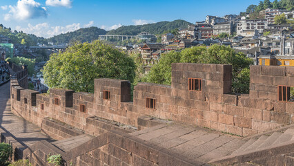 Fototapeta premium The ancient brick city fortress wall. Stone staircase and footpath. Buildings on a hill. A car bridge in the distance. Mountains against the blue sky. China. Fenghuang Ancient Town. Phoenix. 
