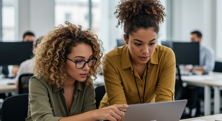 Two women collaborate intently on a laptop in a modern office setting, showcasing focused teamwork and a productive atmosphere.
