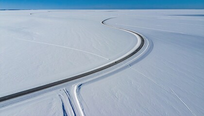 Aerial view of a dark road curving through a vast, snowy plain under a clear blue sky, with slight vehicle visibility