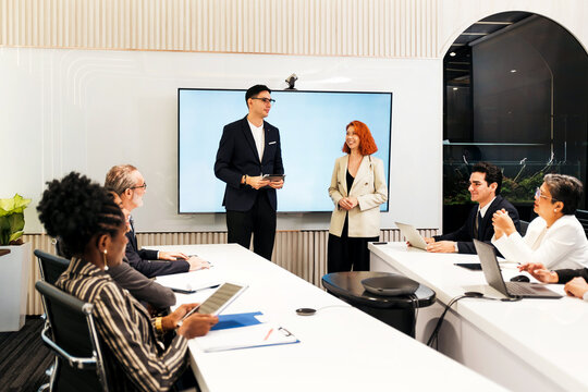 Diverse team in a modern conference room during a presentation. Two colleagues stand by a large screen while others sit with laptops and notes in a corporate meeting.