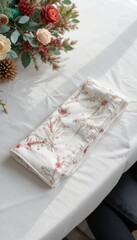 Folded white fabric pouch with floral pattern resting on a textured white surface beside a floral arrangement