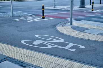 A close-up shot of a white bicycle symbol painted on a dark grey asphalt bike path. The path curves slightly, bordered by a yellow tactile pavement and a pedestrian crossing.