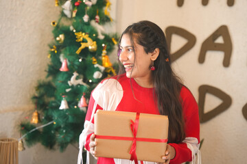 Young happy indian girl holding a wrapped Christmas gift box in front of a decorated tree at home....