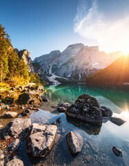 Serene mountain lake scene, clear turquoise water with sun glare, rocky shore, mountain backdrop, autumn colors in trees under blue sky