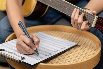 A creative young woman is enjoying her hobby, skillfully playing a guitar while concentrating on writing musical notes in her notebook. The scene captures the essence of music composition and artistic