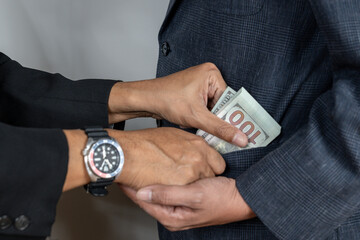An illicit transaction taking place between two business professionals. One man is discreetly slipping a wad of cash into the inner pocket of another person's suit, symbolizing bribery and unethical b