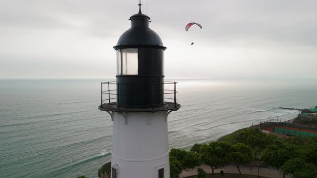 A stunning aerial parallax shot circles the historic Miraflores Lighthouse (Faro La Marina) in Lima, capturing a paraglider soaring over the Pacific Ocean with the beautiful coastline in view