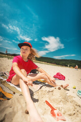 Happy girl in red T-shirt resting on towel, smiling beside small sand sculpture. Bright summer day on beach represents carefree childhood, family vacation and outdoor joy.