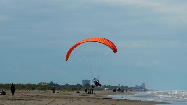 Vibrant Orange Paraglider Descending Gracefully onto a Sandy Beach with Gentle Waves and People Watching