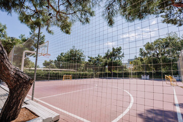 Outdoor basketball and soccer court surrounded by a safety net, trees, and greenery, under a clear blue sky on a sunny day in a public park.