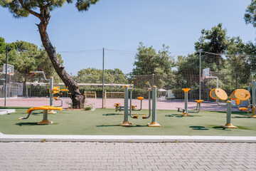 Outdoor gym and fitness equipment in a park near a basketball court, surrounded by trees and safety netting, under a clear blue sky on a sunny day.