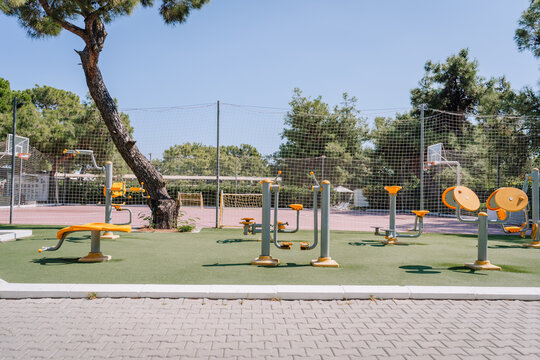 Outdoor gym and fitness equipment in a park near a basketball court, surrounded by trees and safety netting, under a clear blue sky on a sunny day. - Powered by Adobe