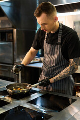Young adult Caucasian man with tattooed arm and short hair wearing apron and gloves pouring oil into frying pan, while cooking food on stove in professional kitchen