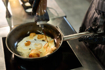 Caucasian middle aged man wearing black gloves preparing scallops in frying pan, pouring sauce over seafood while cooking on stovetop in professional kitchen environment