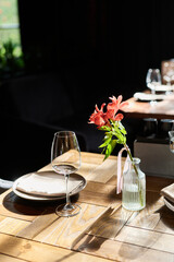 Closeup showing glass vase with pink lily flower standing on wooden restaurant table next to empty wine glass and plate, sunlight casting shadows across surface, background out of focus