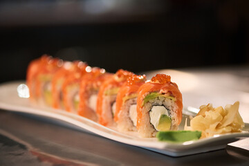 Closeup showing row of sushi rolls with salmon, avocado, rice, and cream cheese arranged on rectangular plate, garnished with pickled ginger and wasabi, no people visible