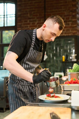 Caucasian young adult man wearing apron and gloves preparing dish in professional kitchen, carefully pouring sauce over plated meal, fresh vegetables visible in background