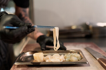 Caucasian young adult man wearing black gloves preparing raw seafood with metal chopsticks in professional kitchen, focusing on arranging ingredients on stainless steel tray
