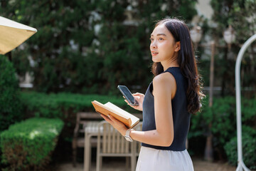 Obraz premium Young woman holding a book and smartphone walking in a garden