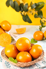 Fresh Persimmons in Wicker Basket with Green Leaves on Yellow Background - Autumn Fruit Still Life