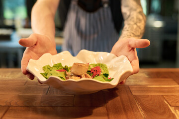 Caucasian young adult man with tattooed arms presenting gourmet dish with leafy greens and meat on decorative plate, standing behind wooden counter in restaurant kitchen