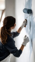A woman is painting a wall with blue paint using a roller, viewed from behind, wearing a long sleeved shirt and gloves