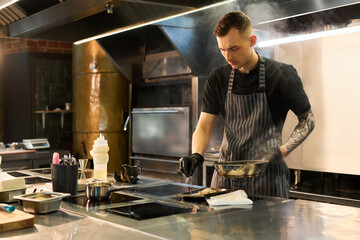 Young adult Caucasian man with tattooed arm cooking food in professional kitchen, holding frying pan and tongs while concentrating on meal preparation at workstation
