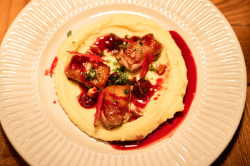 Gourmet dish featuring pieces of cooked meat arranged on creamy mashed potatoes with red berry sauce drizzled on top, served on plate, photographed from above