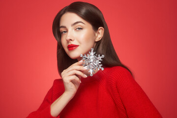 Beautiful young woman with red lipstick, shiny clean skin and face, showing glowing snowflake, near face and smiling, isolated on a red background. Christmas holidays concept