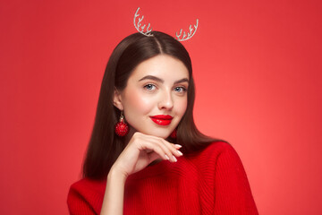 Portrait of beautiful young woman on red background, red lipstick and festive makeup, Christmas toy earrings, shiny glowing skin, Winter holidays concept