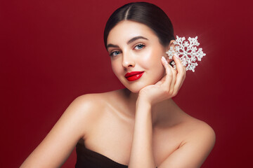 Beautiful young woman with red lipstick, shiny clean skin and face, showing glowing snowflake, near face and smiling, isolated on a red background. Christmas holidays concept