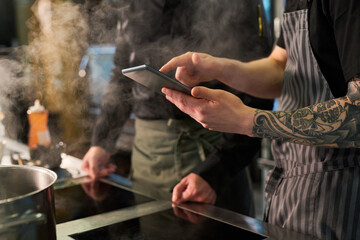 Young adult Caucasian man with tattooed arm using digital tablet in professional kitchen while another young adult man standing nearby, both engaged in culinary preparation near stove