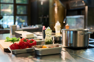 Fresh vegetables including red bell peppers and cherry tomatoes arranged on metal trays near stainless steel pot boiling on stove, in professional kitchen with blurred background
