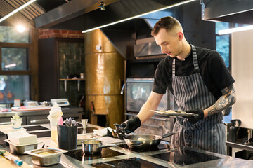 Caucasian young adult man with tattooed arm preparing food in professional kitchen, wearing apron and gloves, focused on cooking process with various utensils and ingredients visible