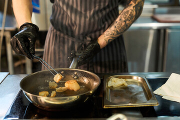 Tattooed young adult Caucasian man wearing black gloves searing scallops in frying pan on stovetop, using tongs to turn seafood, stainless steel tray with ingredients beside him