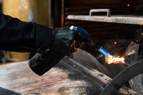 Caucasian middle aged man wearing black gloves using blowtorch to heat metal inside industrial workshop or commercial kitchen, sparks flying as intense blue flame contacts surface, close up of hands