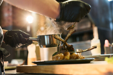Caucasian young adult man wearing black gloves plating cooked lamb chops and vegetables in restaurant kitchen, holding saucepan and spoon while adding sauce to dish, steam rising from food © pressmaster