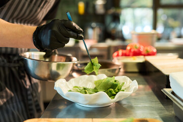 Caucasian young adult man wearing black gloves preparing fresh salad in professional kitchen, holding metal bowl and using tongs to place lettuce leaves into dish
