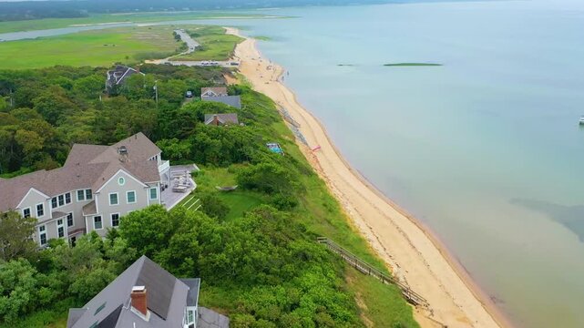 Panoramic drone perspective of Cape Cod shoreline at high tide, highlighting rows of beach houses facing the Atlantic, where a boat drifts offshore against a moody horizon.