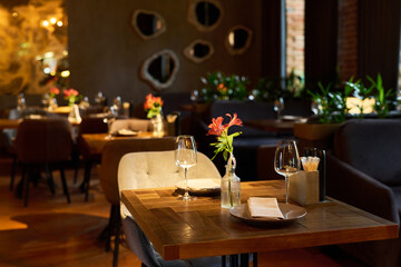 Empty restaurant interior showing wooden table set with plates, wine glasses, napkins, and fresh flowers, modern dining area arranged for guests, soft lighting illuminating space